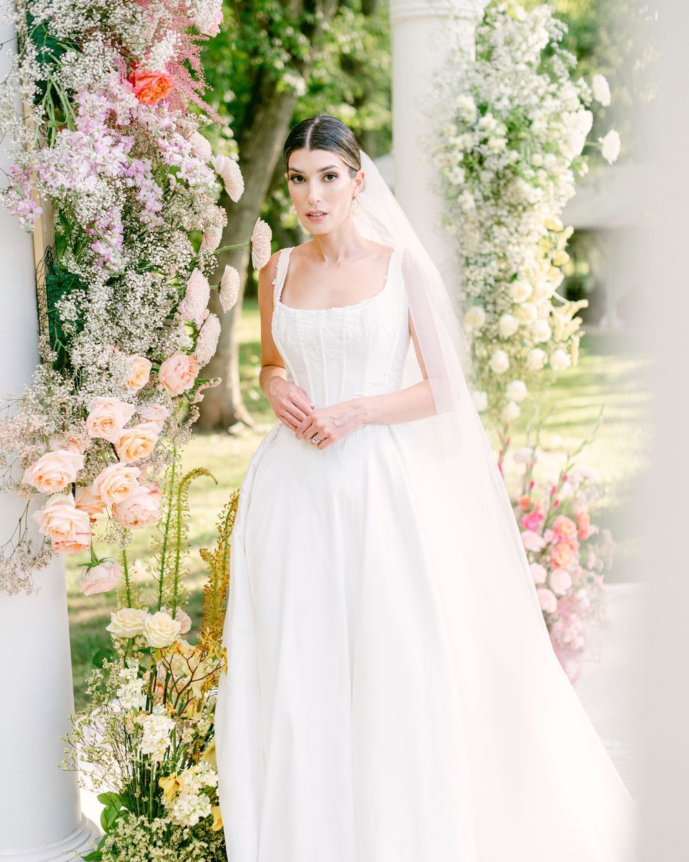 A bride in a classic white wedding gown stands elegantly among floral arrangements and columns in a garden setting.