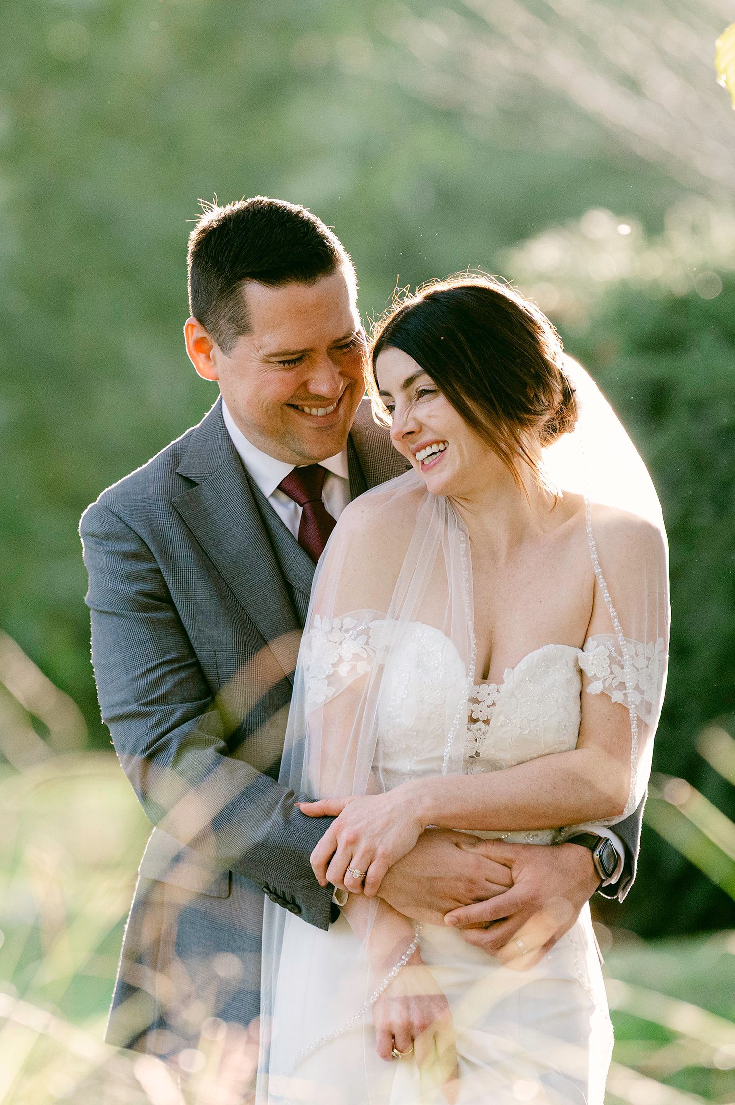 A romantic wedding couple embraces and laughs together in a sunlit outdoor setting wearing formal attire.