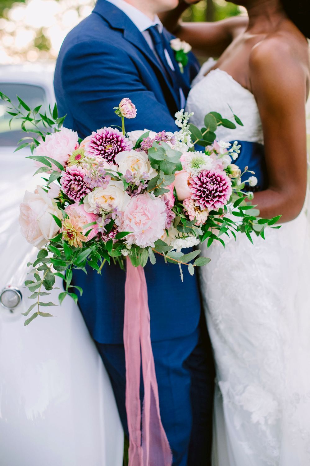 Wedding party members in navy suits and white dresses hold pink and purple floral bouquets with flowing ribbons.