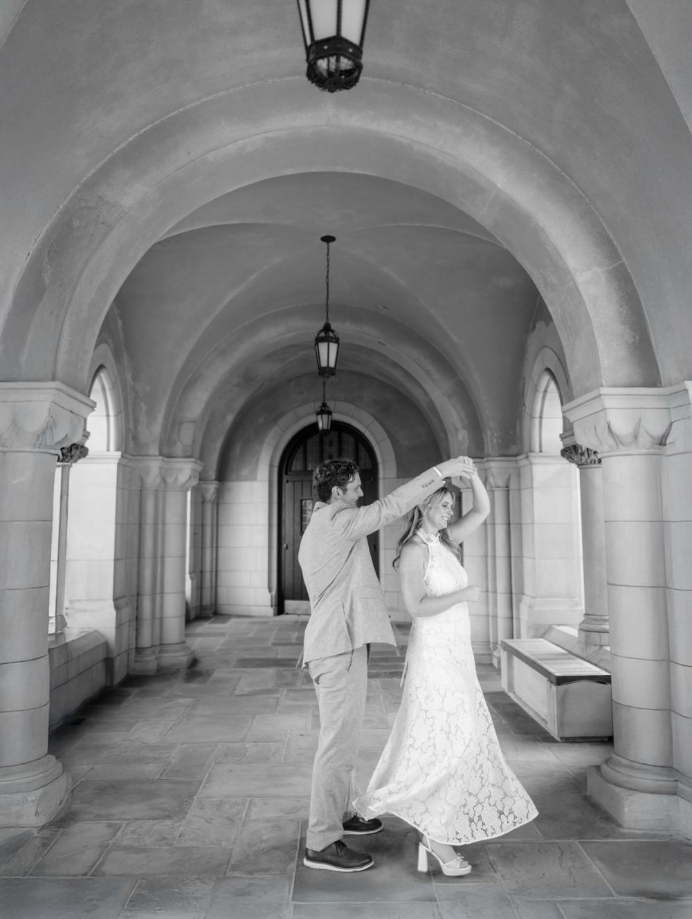 A black and white photo captures a dancing couple in an elegant arched hallway with pendant lights.
