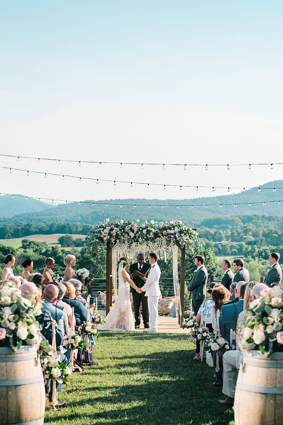 Outdoor wedding ceremony on a hilltop lawn with wine barrels and string lights overlooking mountain views.