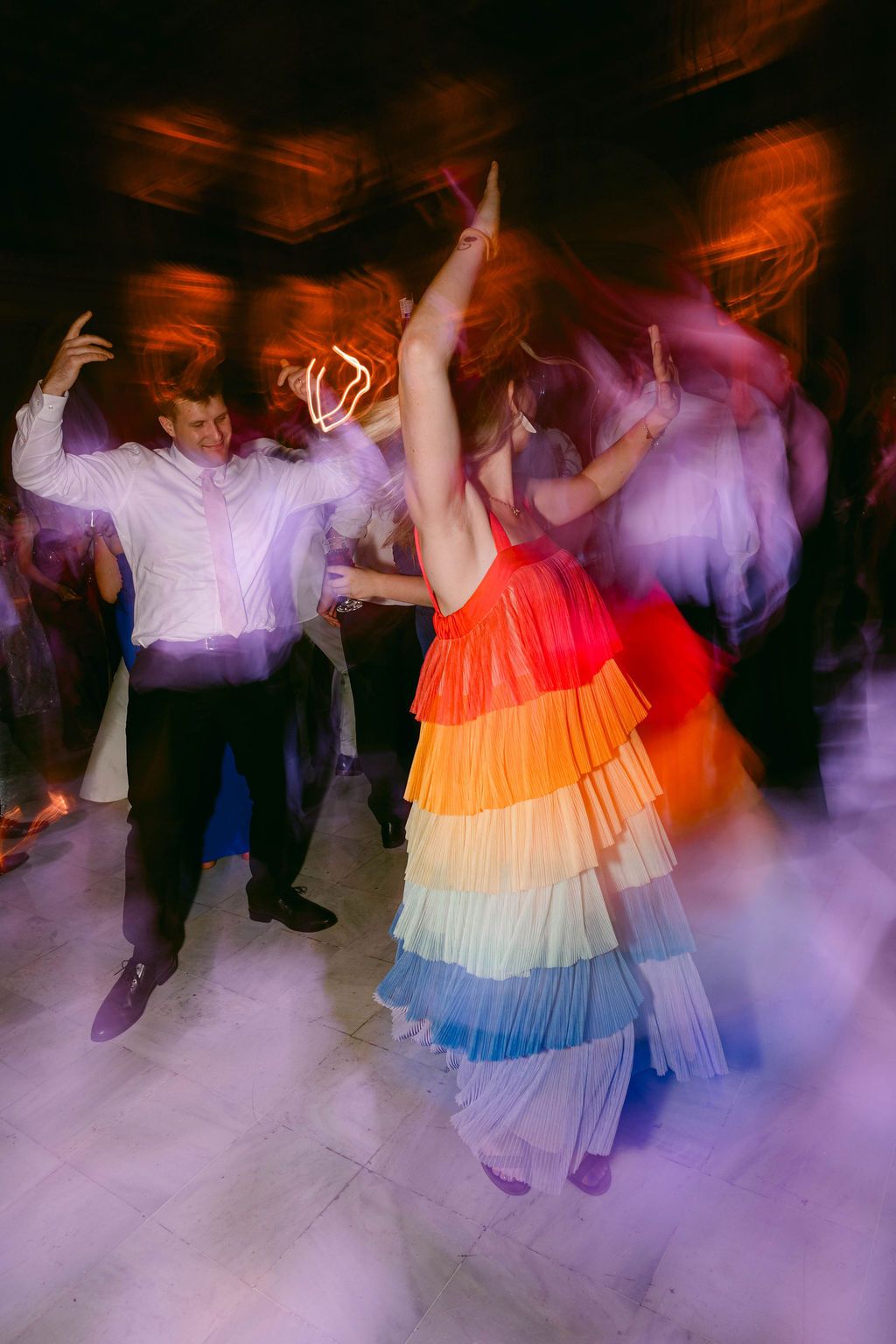 Blurred motion of dancers in colorful dresses moving on a foggy dance floor with purple lighting effects.