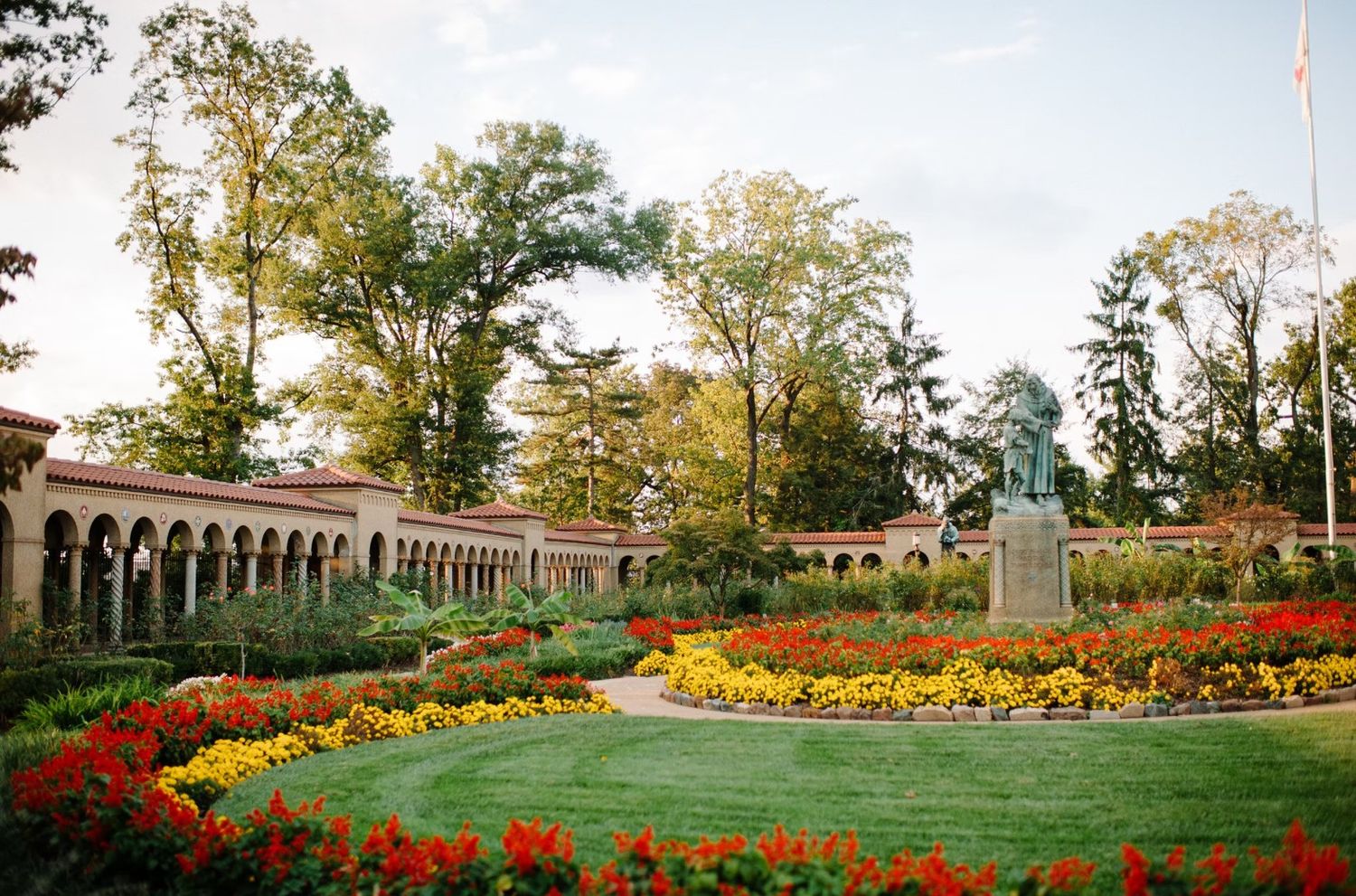 Beautiful garden courtyard with arched colonnade and colorful flower beds featuring red and yellow blooms at sunset.