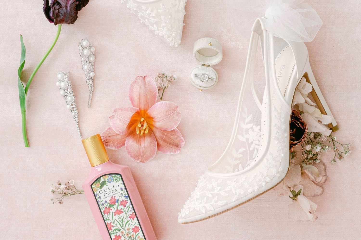 Vintage inspired white wedding accessories including lace shoes, flowers, and delicate decor arranged on pink background.