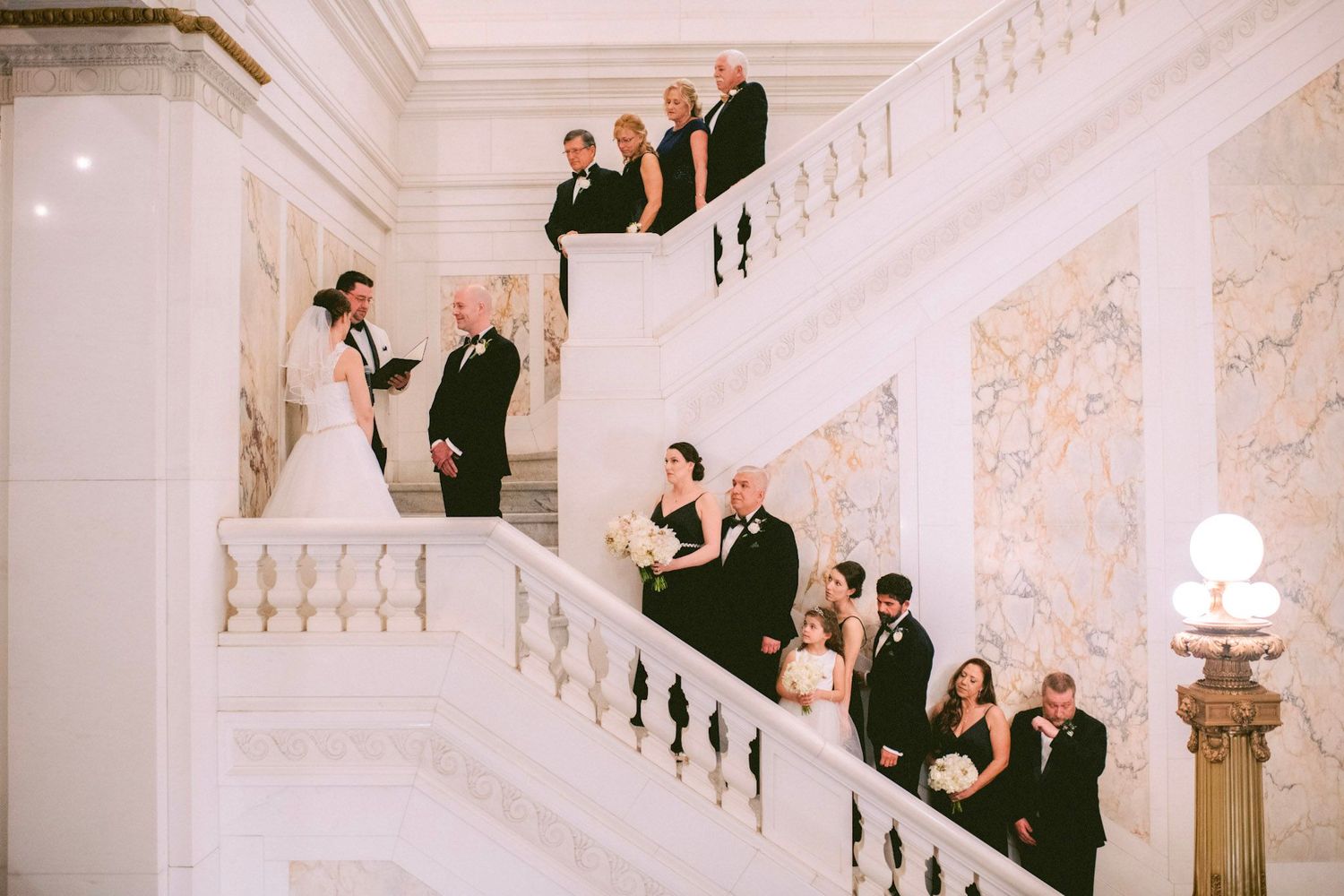 Wedding party members in formal attire descend an elegant white marble staircase during a celebration.