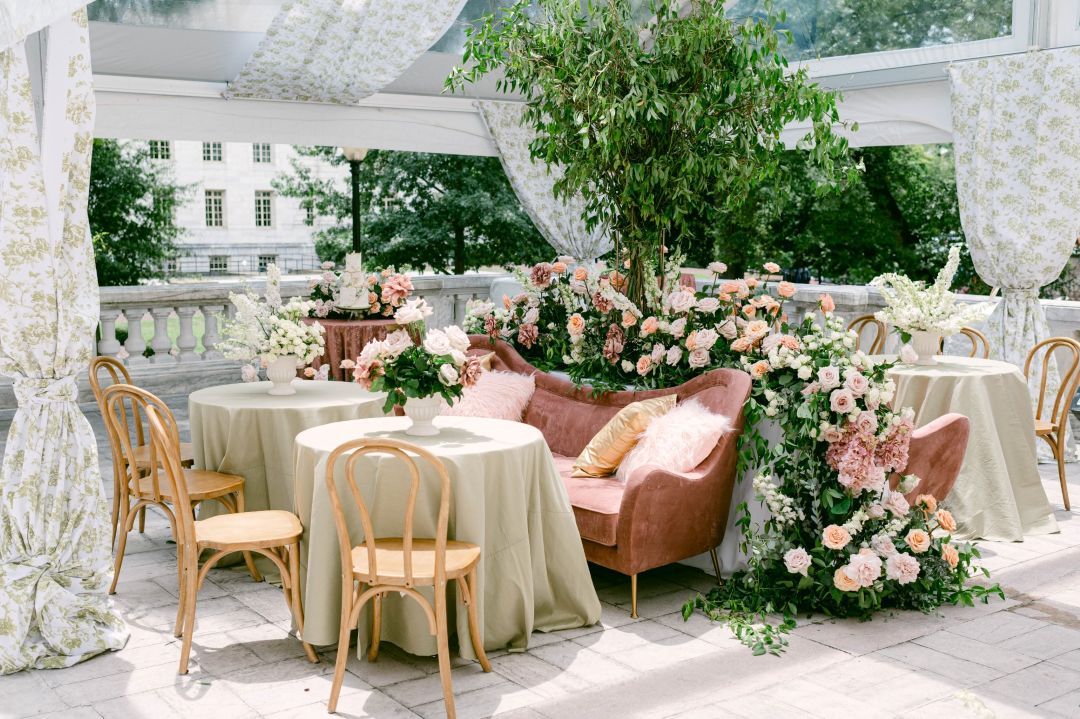 Elegant white and blush pink wedding reception decor with floral arrangements and soft draping in a conservatory setting.