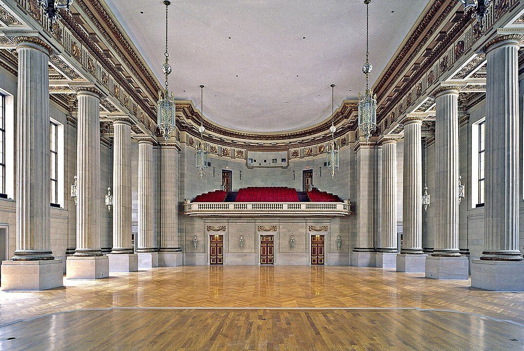Elegant neoclassical ballroom interior with white columns, wooden floors, and red-upholstered balcony seating.