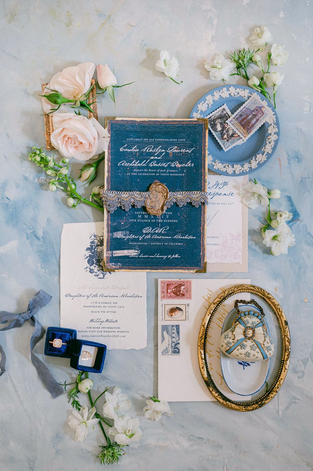 Elegant wedding invitation suite with navy blue and gold details alongside pink roses and vintage decor on marble background.