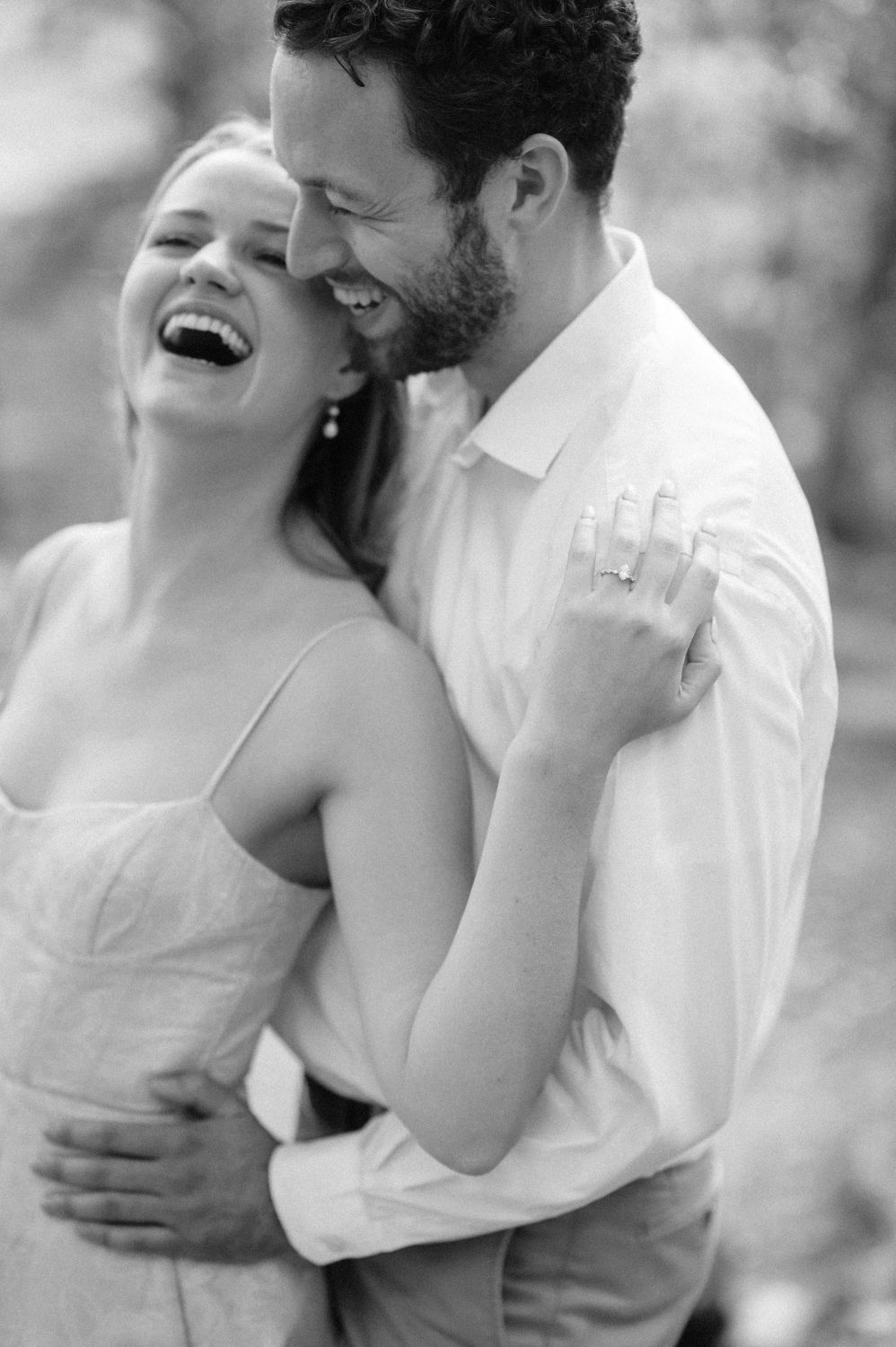 Black and white photo of a laughing couple embracing outdoors, woman in spaghetti strap dress, man in button-up shirt.
