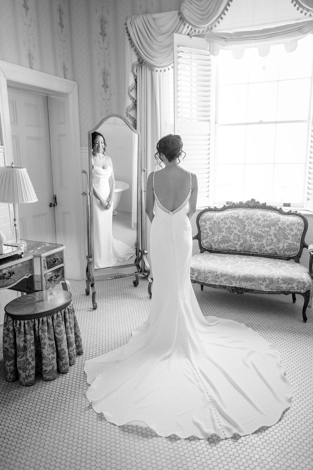 Elegant backlit wedding dress with long train shown in black and white photo as bride looks into full-length mirror in getting ready room.