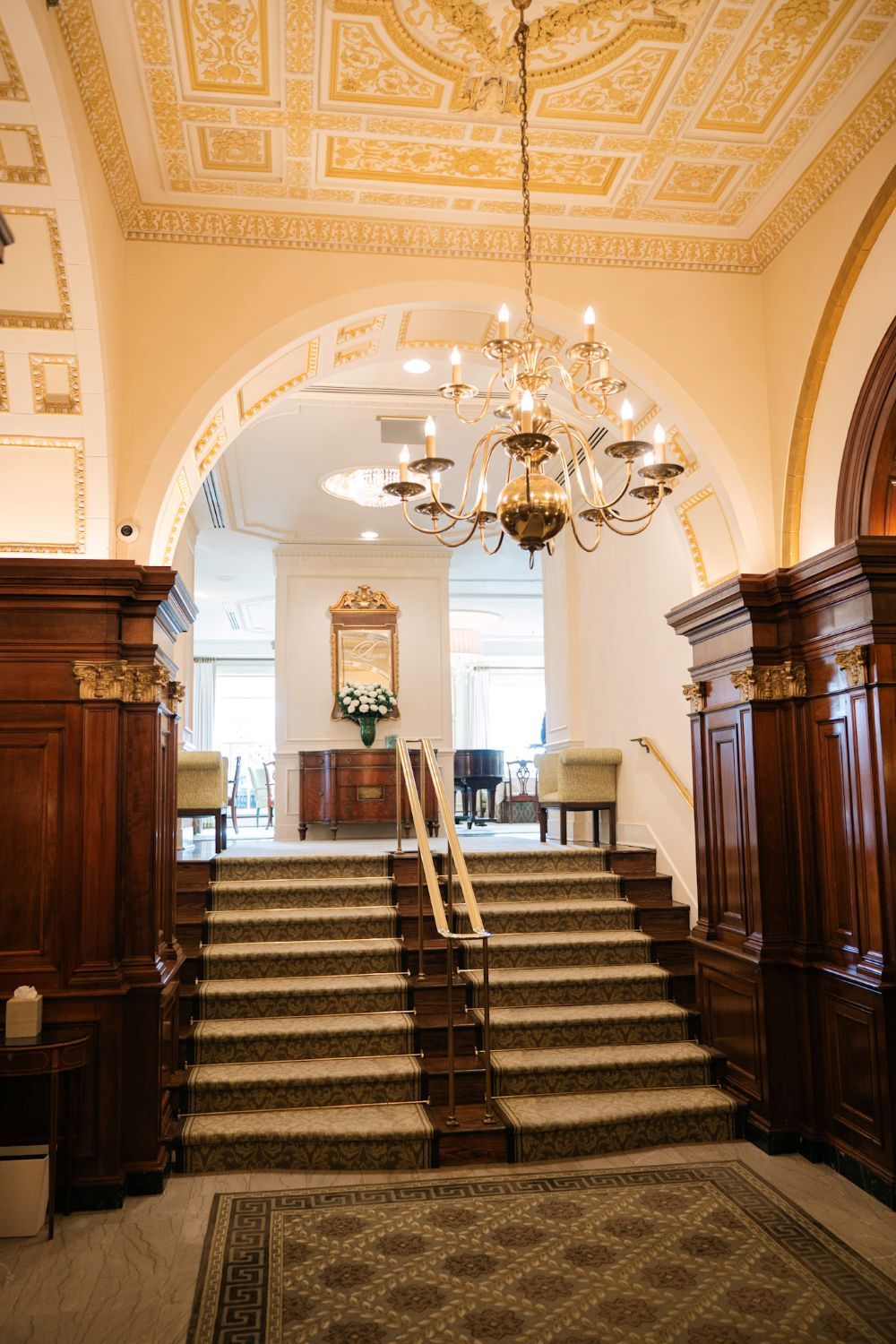 Elegant grand staircase with ornate ceiling and dark wood paneling in historic interior with chandeliers.