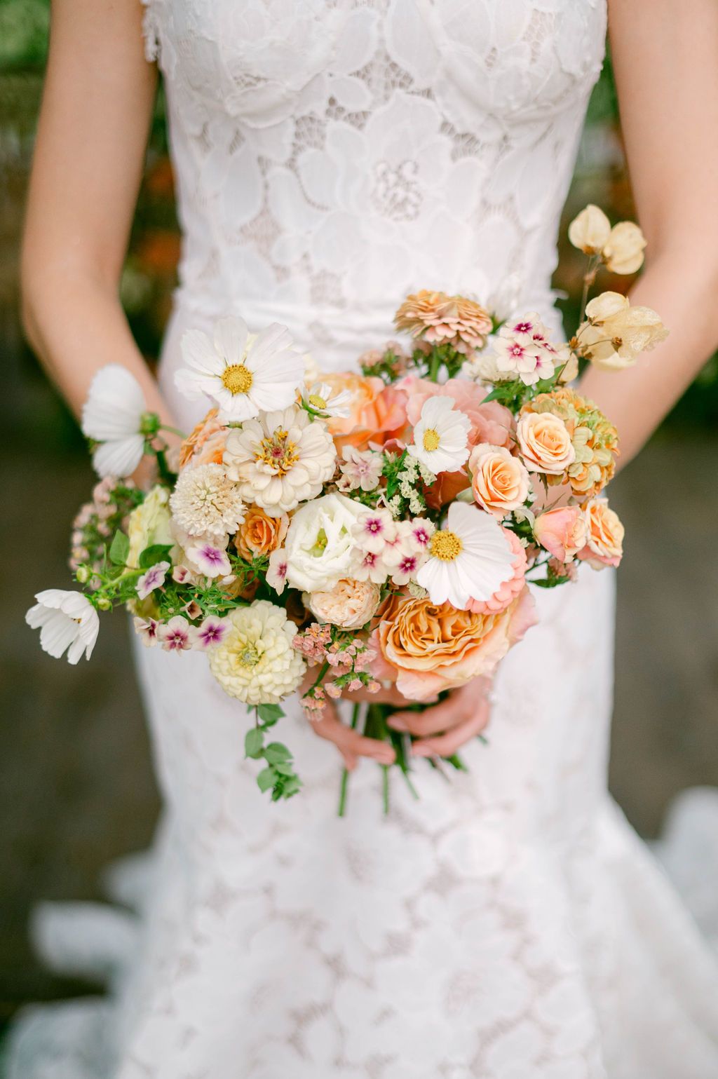 A wedding bouquet in peach and cream tones with roses and mixed flowers held against a white lace dress.
