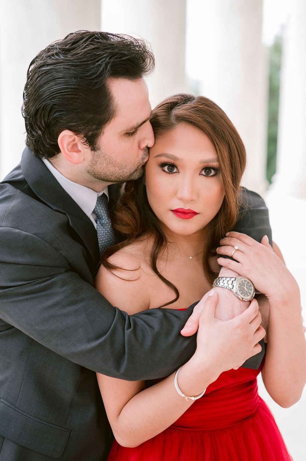 A romantic couple in formal attire embraces intimately, with her wearing a red dress and him in a dark suit.