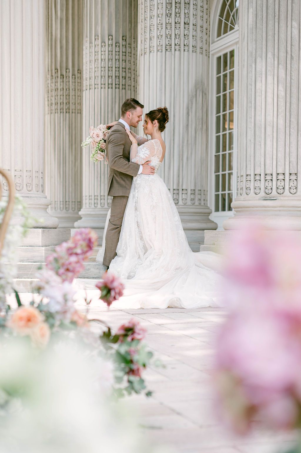 A romantic wedding couple dances in an elegant colonnade with soft pink floral decorations creating a dreamy atmosphere.
