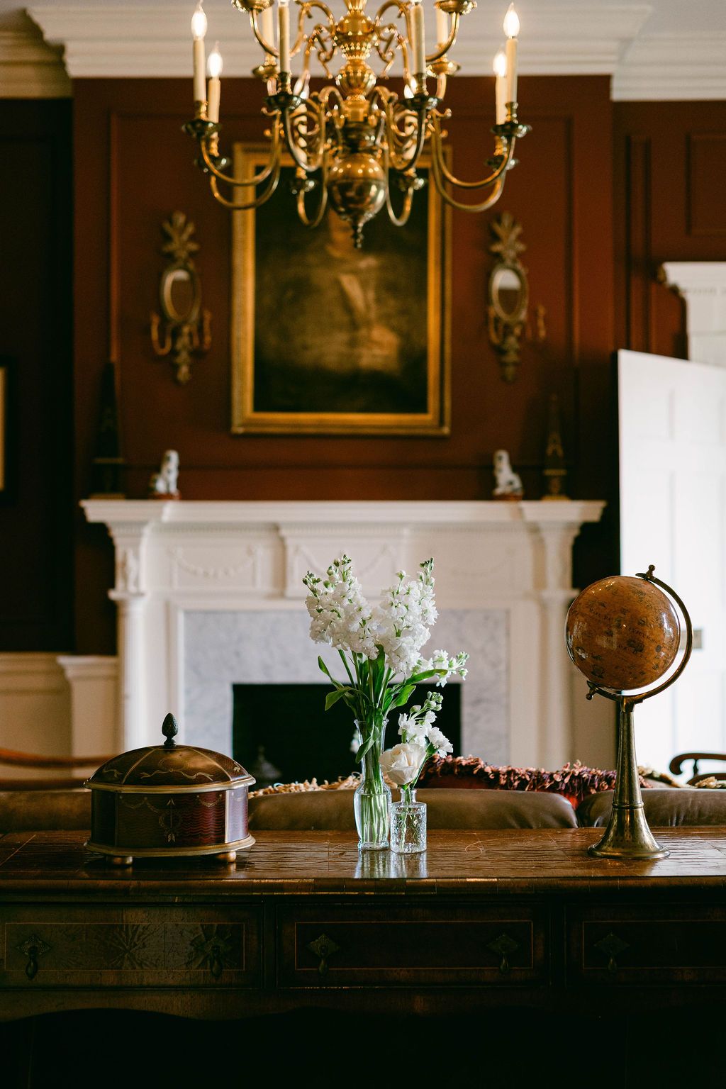 Elegant interior scene with brass chandelier, wood table, and vintage globe silhouetted in moody lighting in front of white fireplace.