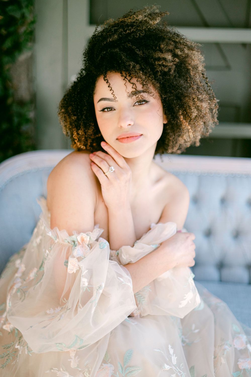 Beautiful portrait featuring natural curly hair and delicate off-shoulder sheer dress while sitting on blue velvet couch.