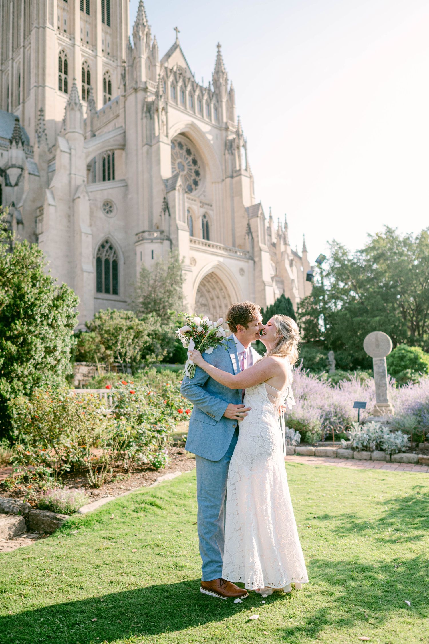 A romantic wedding couple embraces on a sunny day outside a grand Gothic cathedral with manicured gardens.