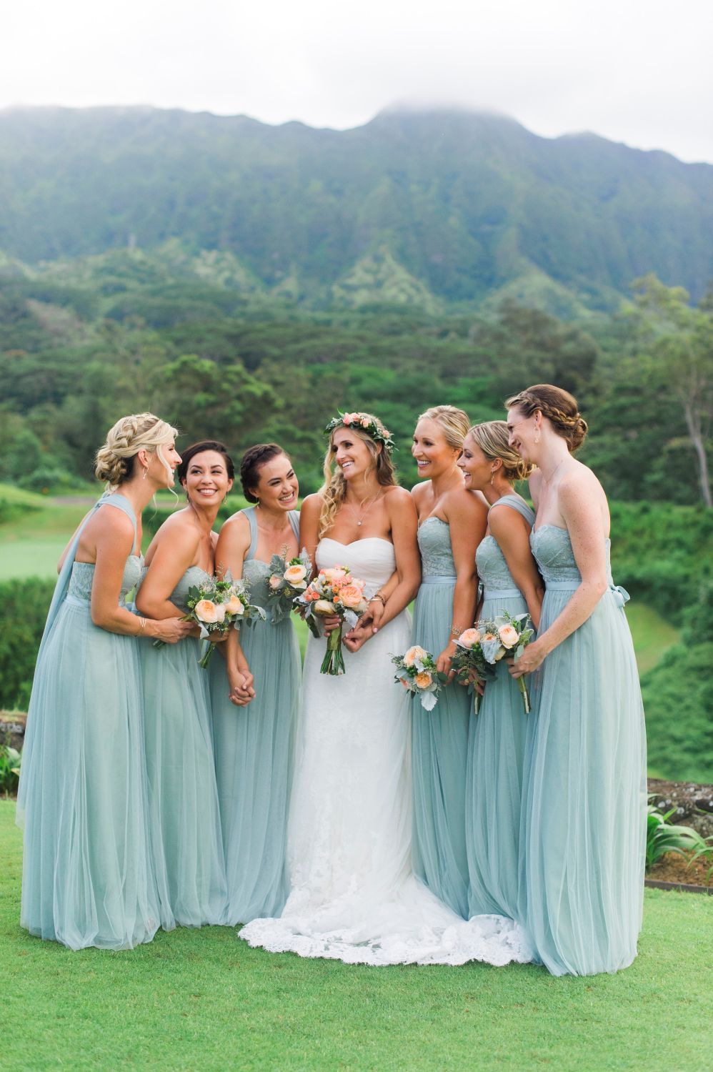 Wedding party in flowing mint green bridesmaid dresses gather on a lush golf course with misty mountains in background.