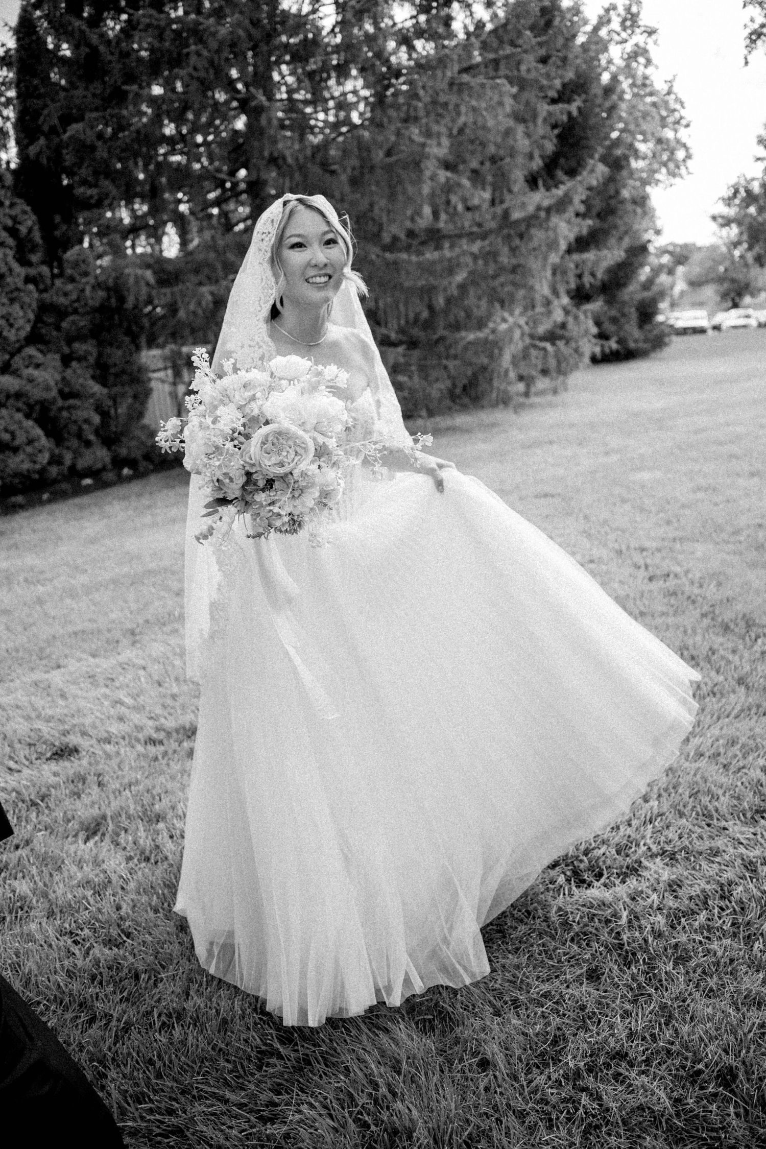 Black and white photo of a smiling bride in a flowing gown and veil holding a bouquet outdoors.