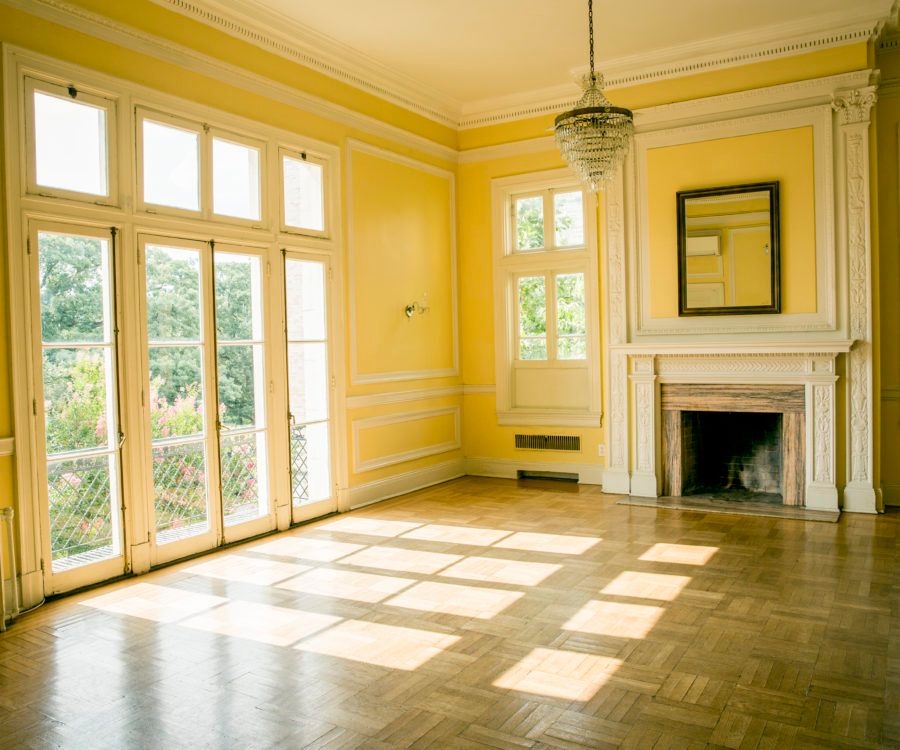 Bright sunlight streams through multiple windows into an empty yellow room with hardwood floors and crown molding.