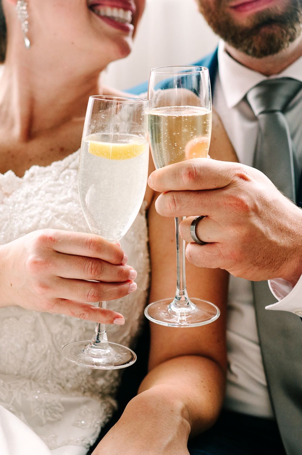 A couple toasts with champagne flutes at a wedding celebration, showing lace dress detail and formal attire.