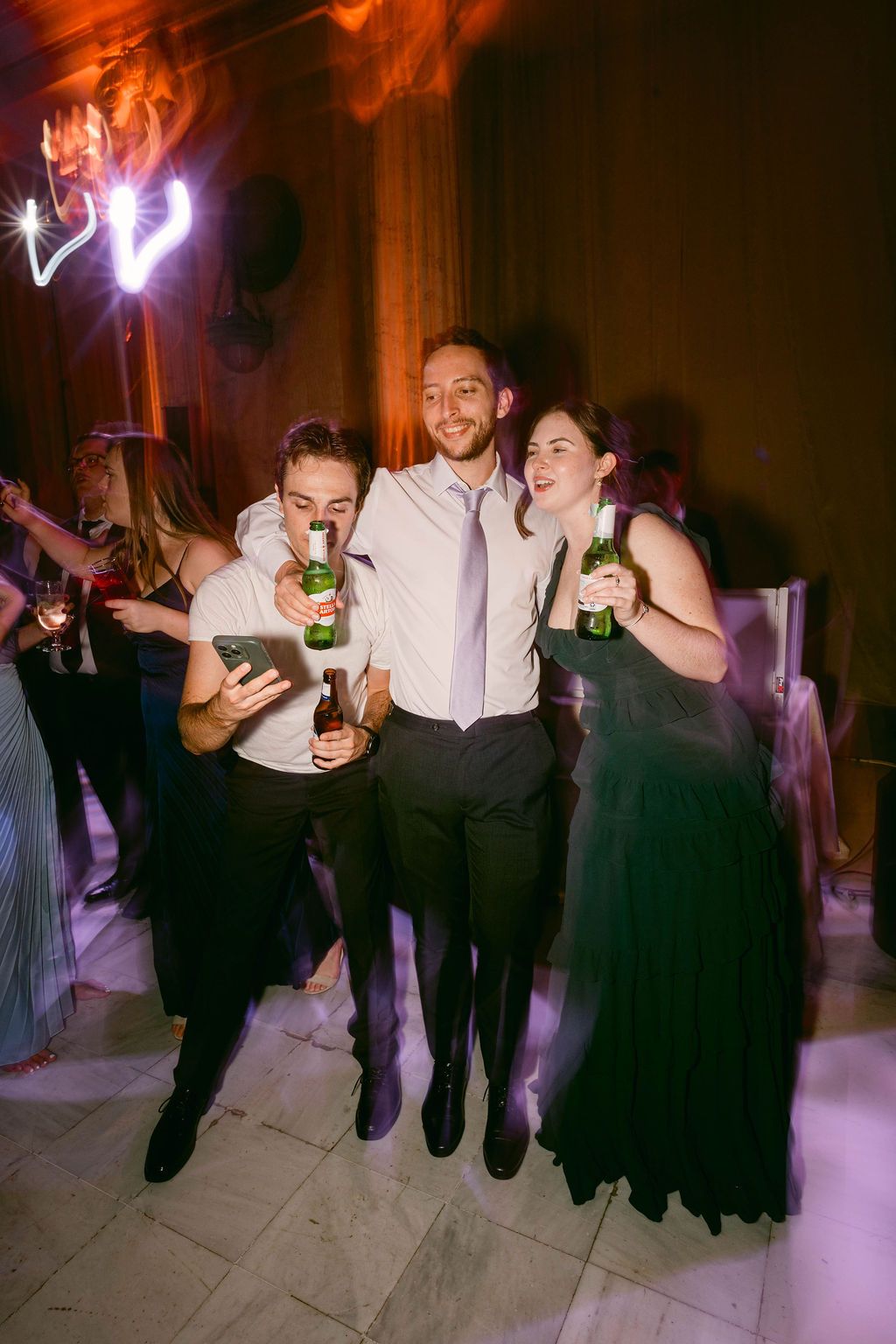 Wedding party guests dance and celebrate with drinks in their hands during a reception under purple lighting.
