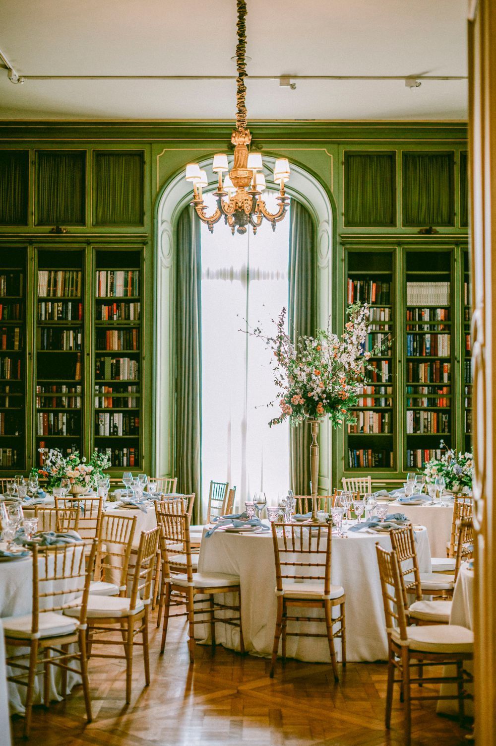 Elegant wedding reception venue with sage green walls, built-in bookshelves, crystal chandeliers and round dining tables.