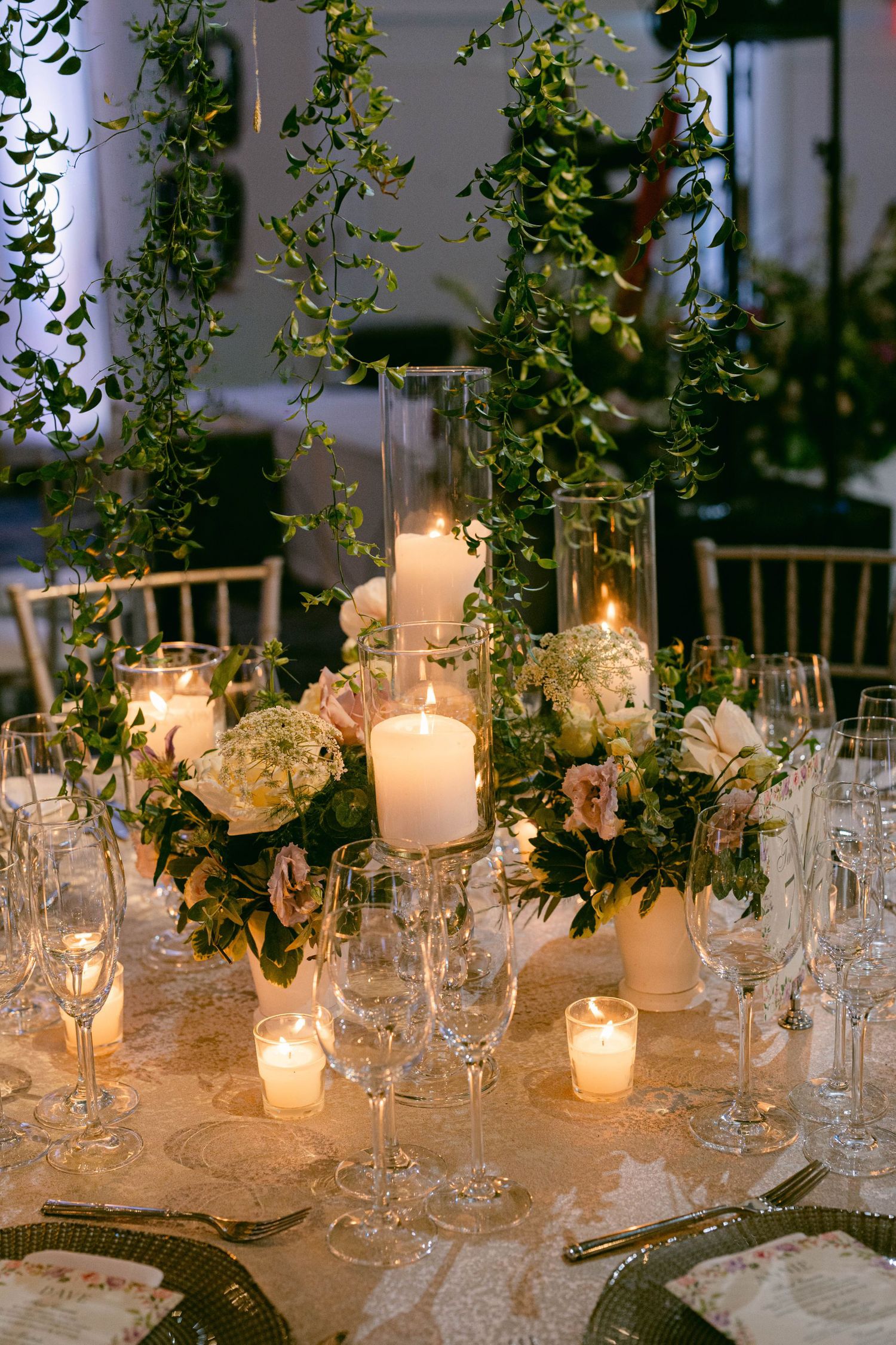 Elegant wedding reception table with white pillar candles, blush floral arrangements, crystal glassware, and cascading greenery centerpiece.
