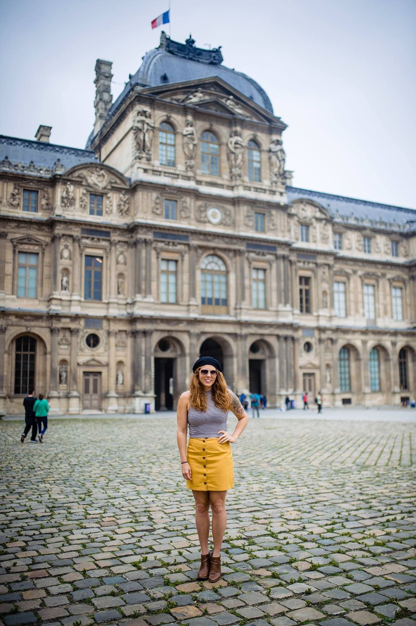 A tourist in a yellow skirt and black beret poses in front of the historic Louvre Museum in Paris on a cloudy day.
