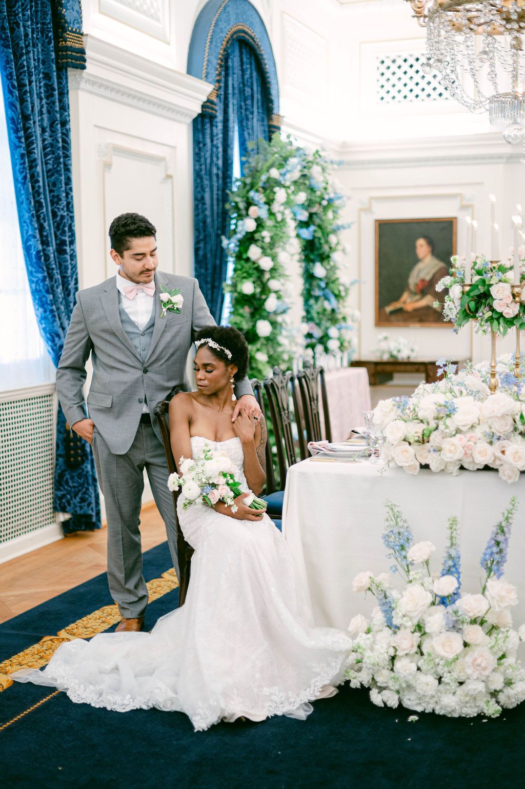 Elegant wedding reception setup with blue velvet curtains, white floral arrangements and luxurious table settings in a grand ballroom.