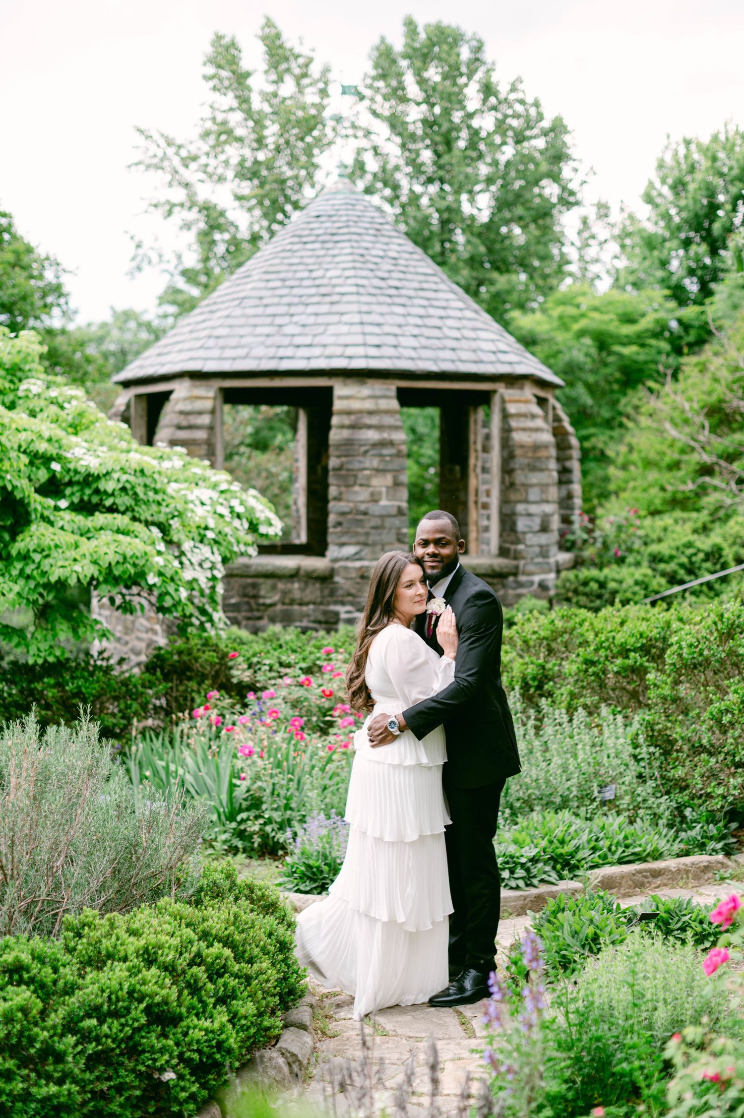 Couple embracing in lush garden with stone gazebo, woman in white dress and man in black suit.