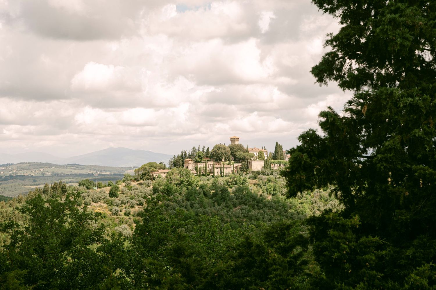 Rolling hills and medieval architecture of Tuscany stretch across a panoramic countryside landscape under cloudy skies.