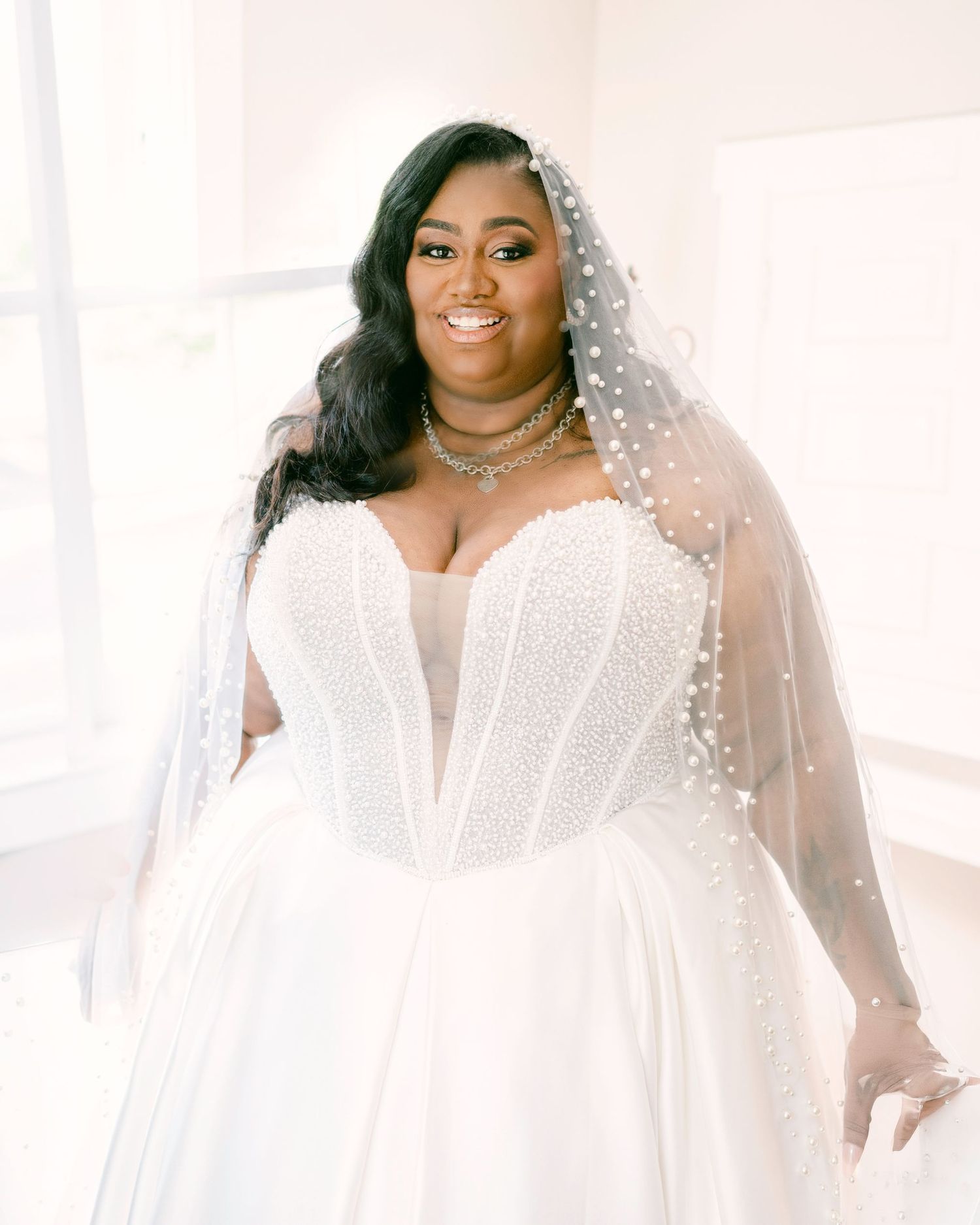 A radiant plus-size bride in a pearl-embellished strapless white ballgown with a pearl-dotted veil and layered necklace.