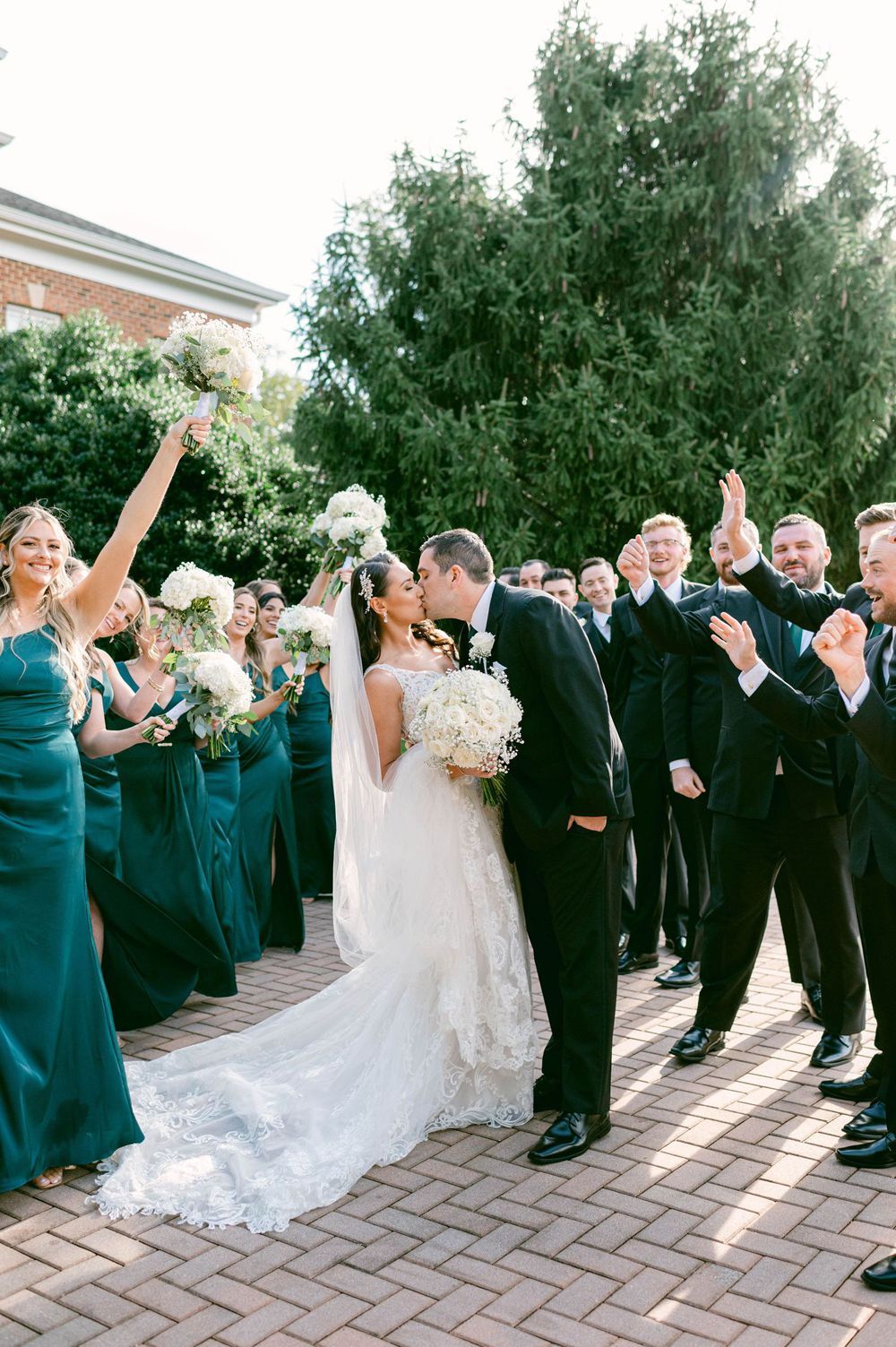 Wedding party celebrates joyfully on brick patio with bridal party in teal dresses and groomsmen in dark suits.