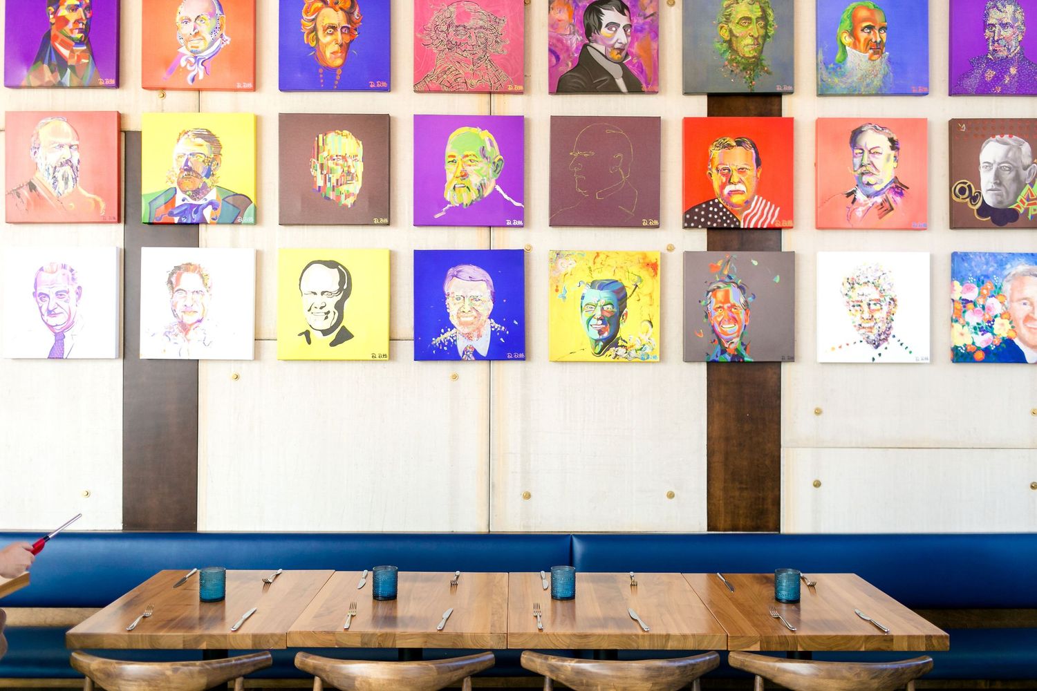 Colorful pop art style portraits hang above a wooden bench with blue metal backing in a restaurant or cafe setting.