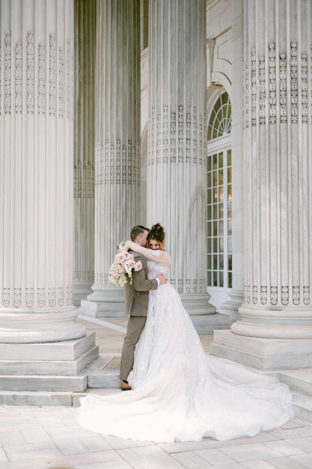 Elegant wedding couple in formal attire embraces against white marble columns in classic architectural setting.