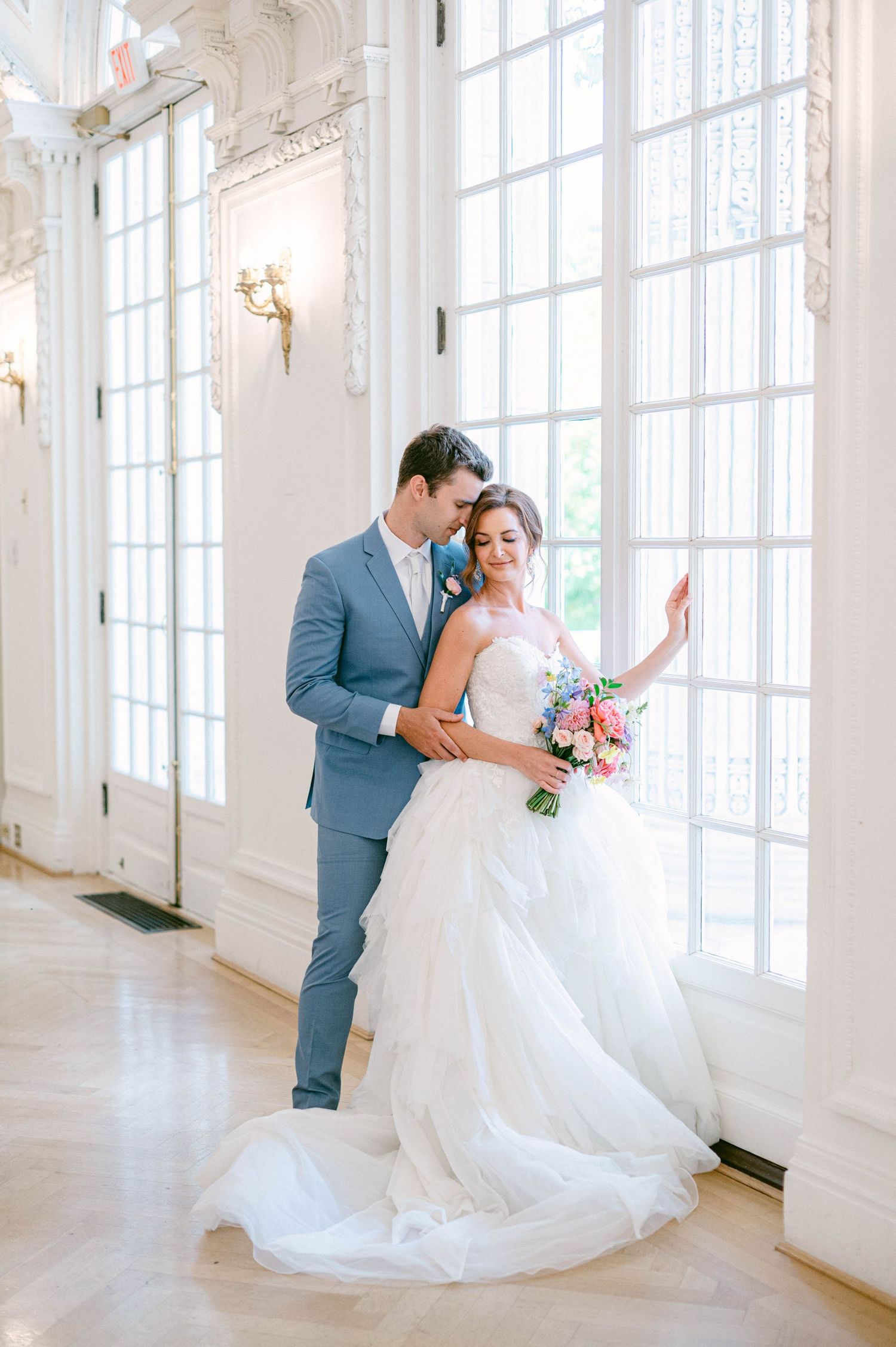 Bride and groom share a romantic moment in an elegant white hall, she holds a colorful bouquet in a flowing tulle gown.