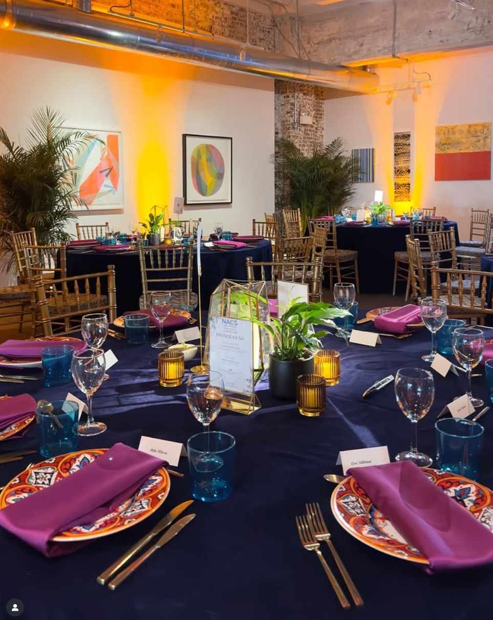 Formal dining table set with navy tablecloth, purple napkins, and colorful charger plates in an industrial-style event space.