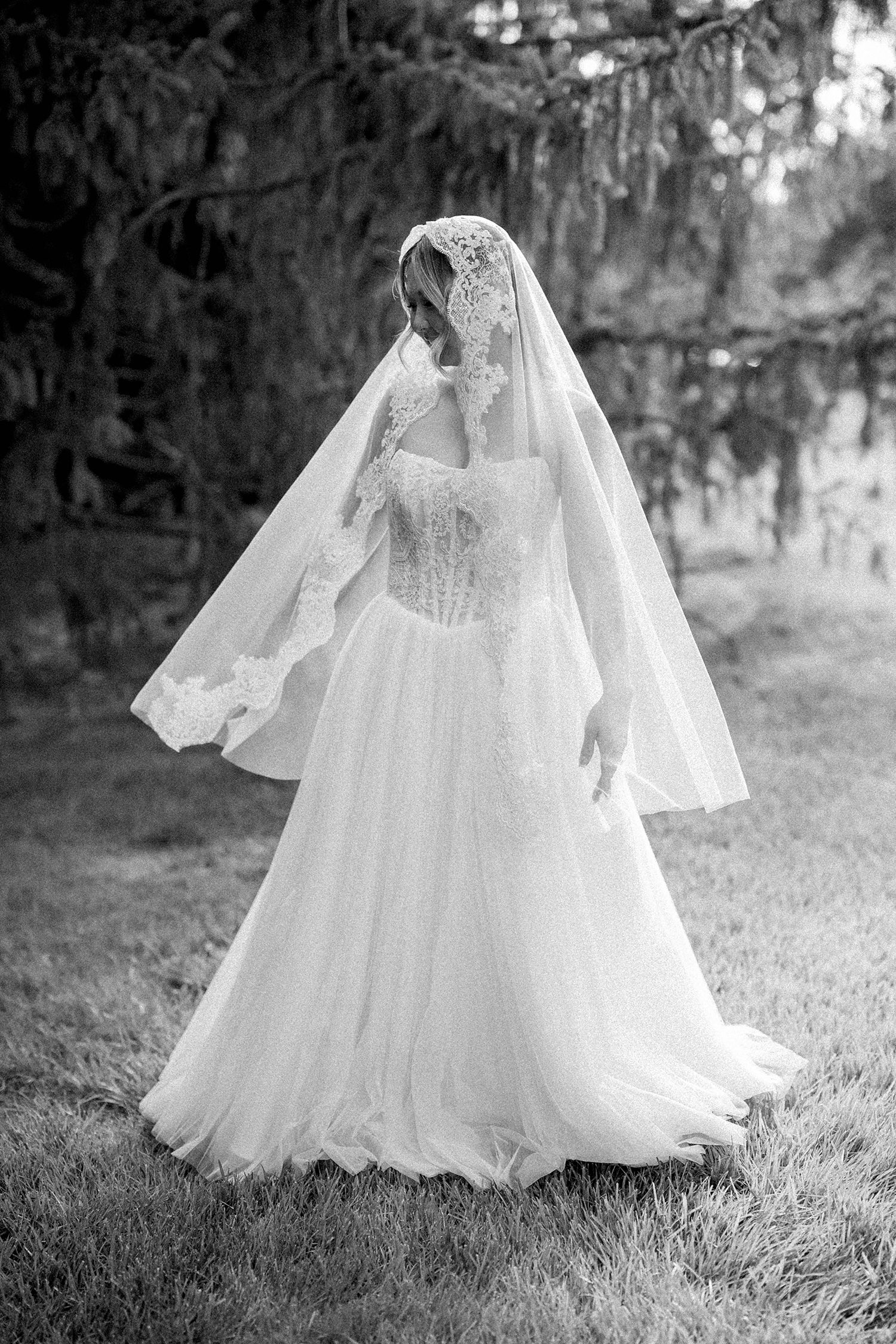 Black and white photo of a bride in a lace wedding gown and veil standing outdoors under a tree.