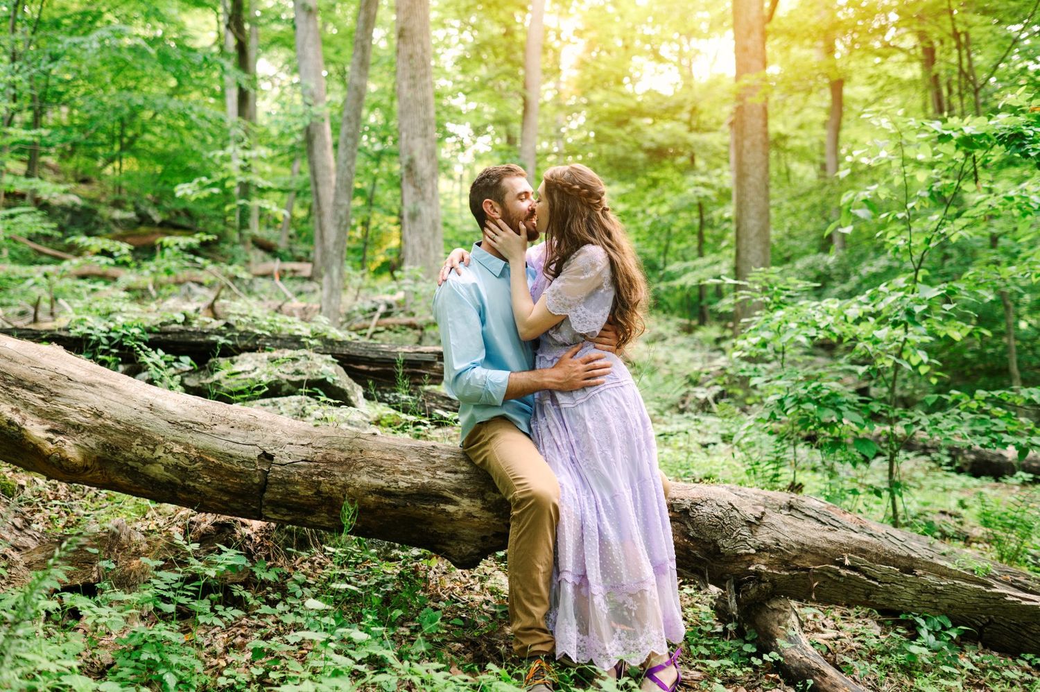 Couple sharing a romantic kiss on a fallen log in a lush green forest during a dreamy outdoor engagement session.