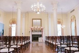 Elegant wedding ceremony venue with rows of dark wooden chairs, crystal chandelier, and white fireplace mantel at the altar.