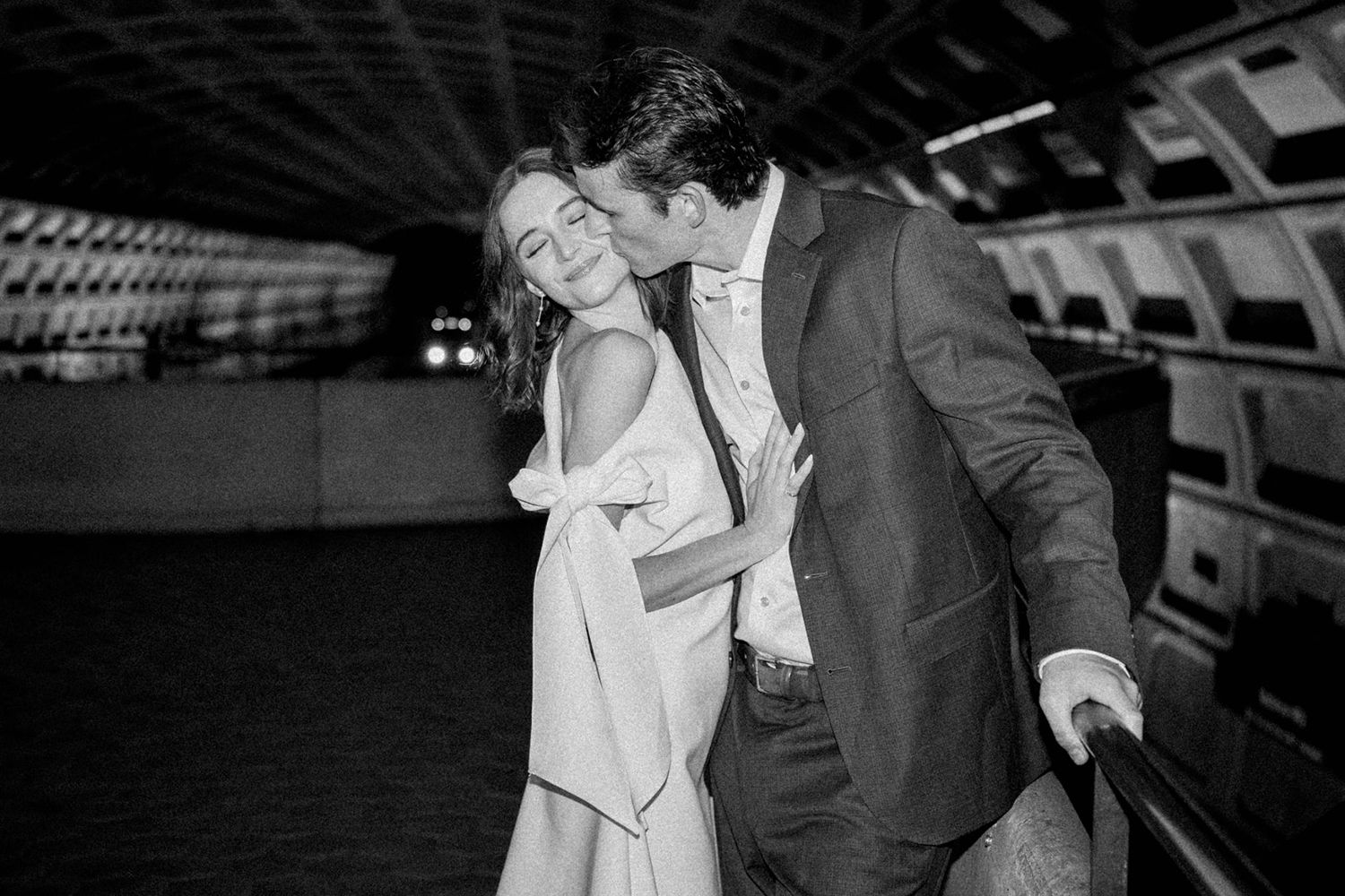 Black and white photo of a couple sharing a romantic kiss at a subway station, woman in elegant dress.