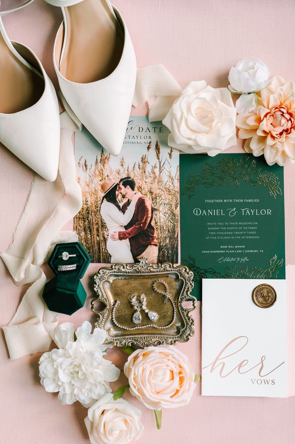 Elegant wedding flat lay with green invitation, save the date, white heels, ring box, earrings, and blush florals on pink background.