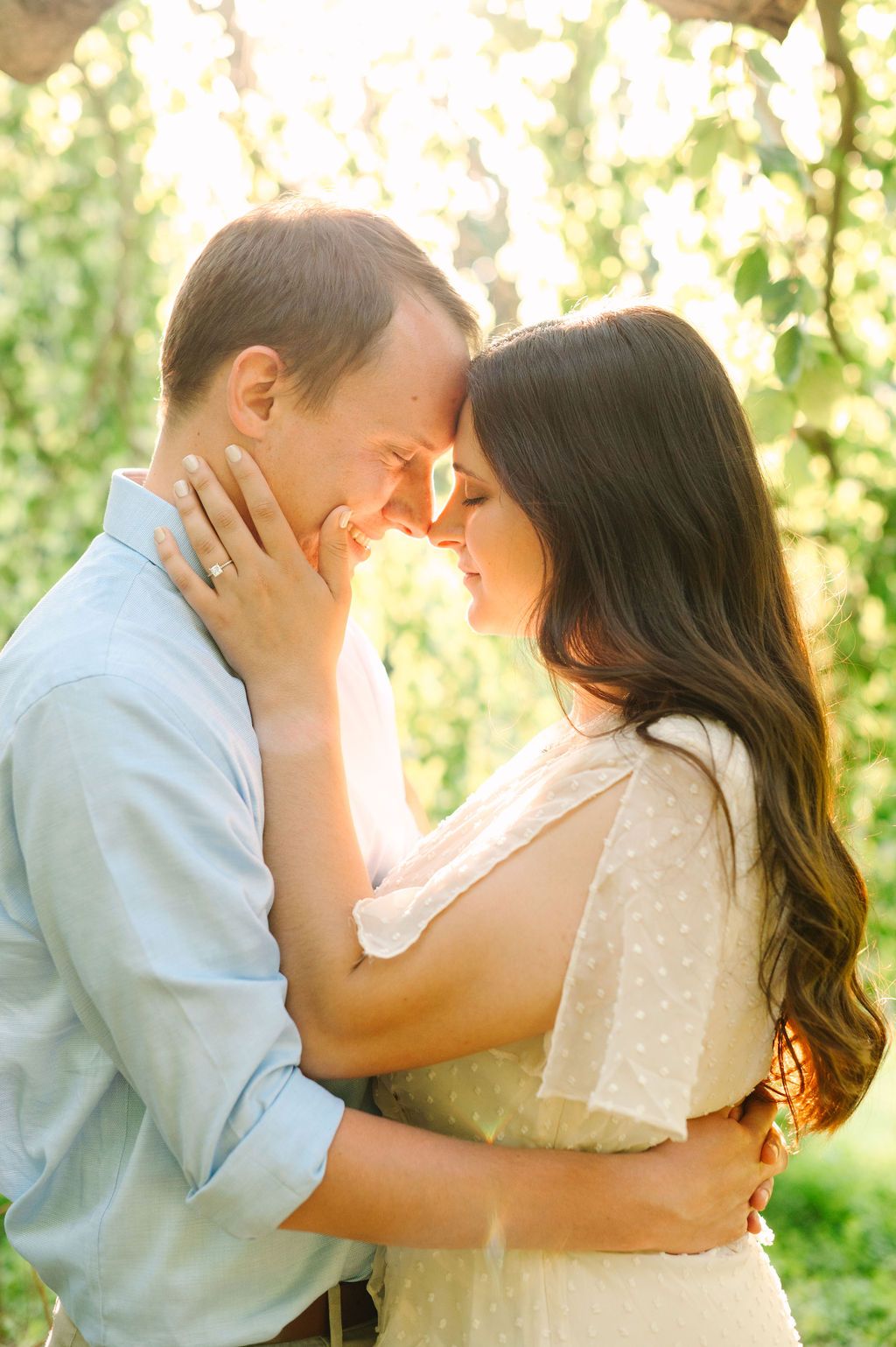 Couple sharing a romantic forehead touch outdoors, bathed in golden sunlight among lush green trees.