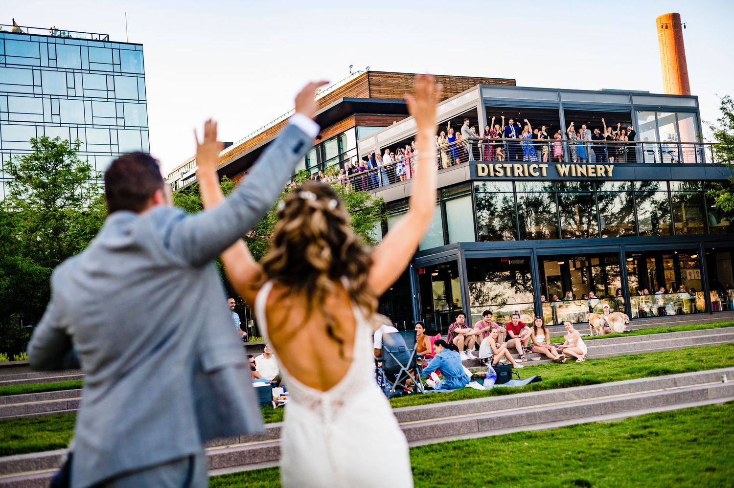 A wedding couple raises their arms in celebration outside District Winery in Washington DC with spectators watching from balcony above.