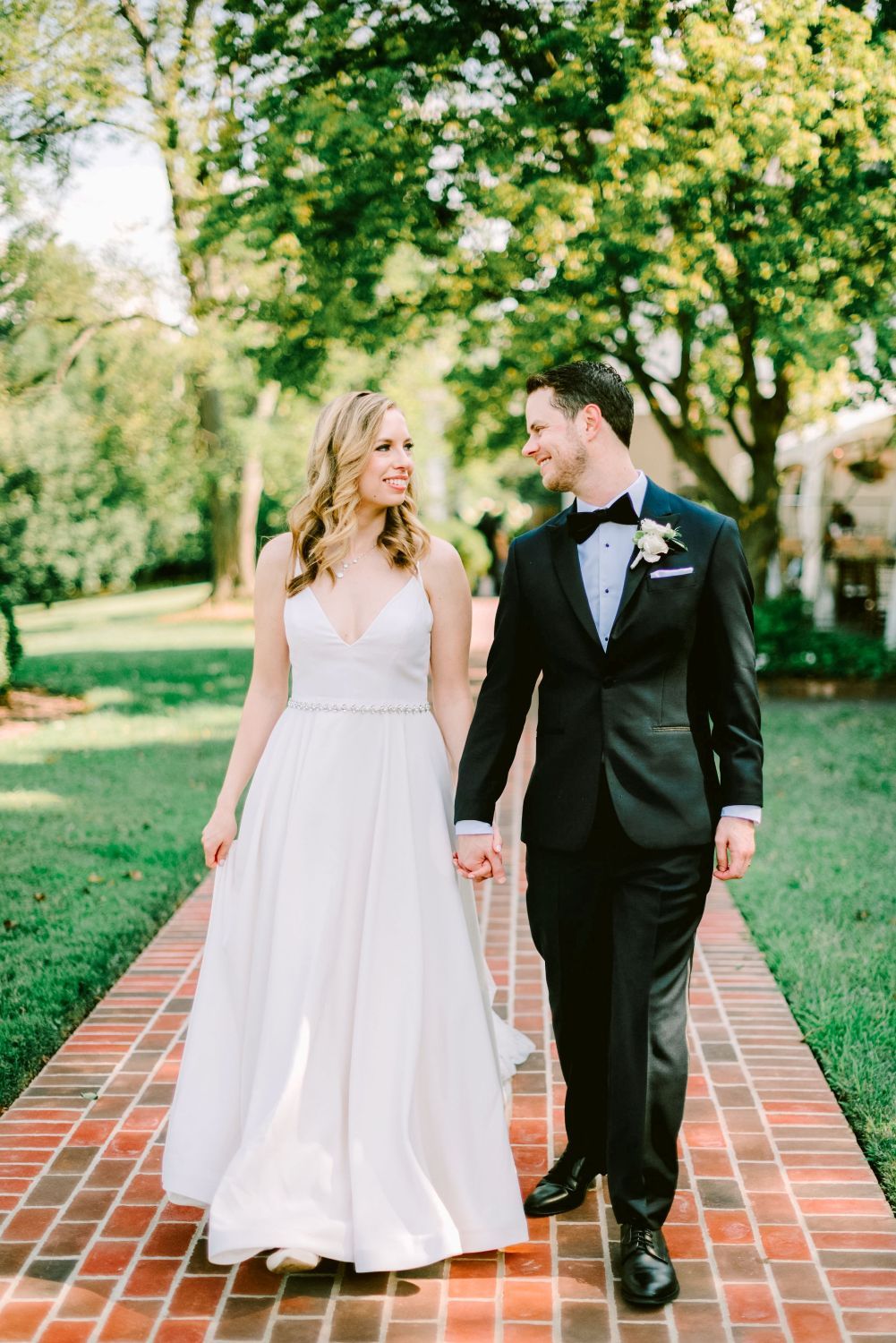 A couple in wedding attire walks together on a brick path surrounded by lush green trees and gardens.