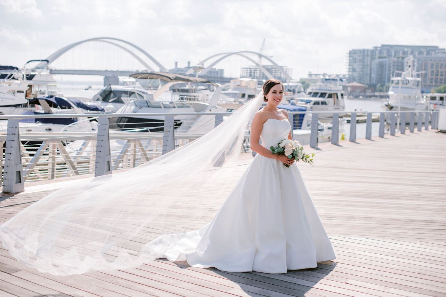 A person in an elegant white wedding gown twirls on a wooden dock with boats and a marina in the background.