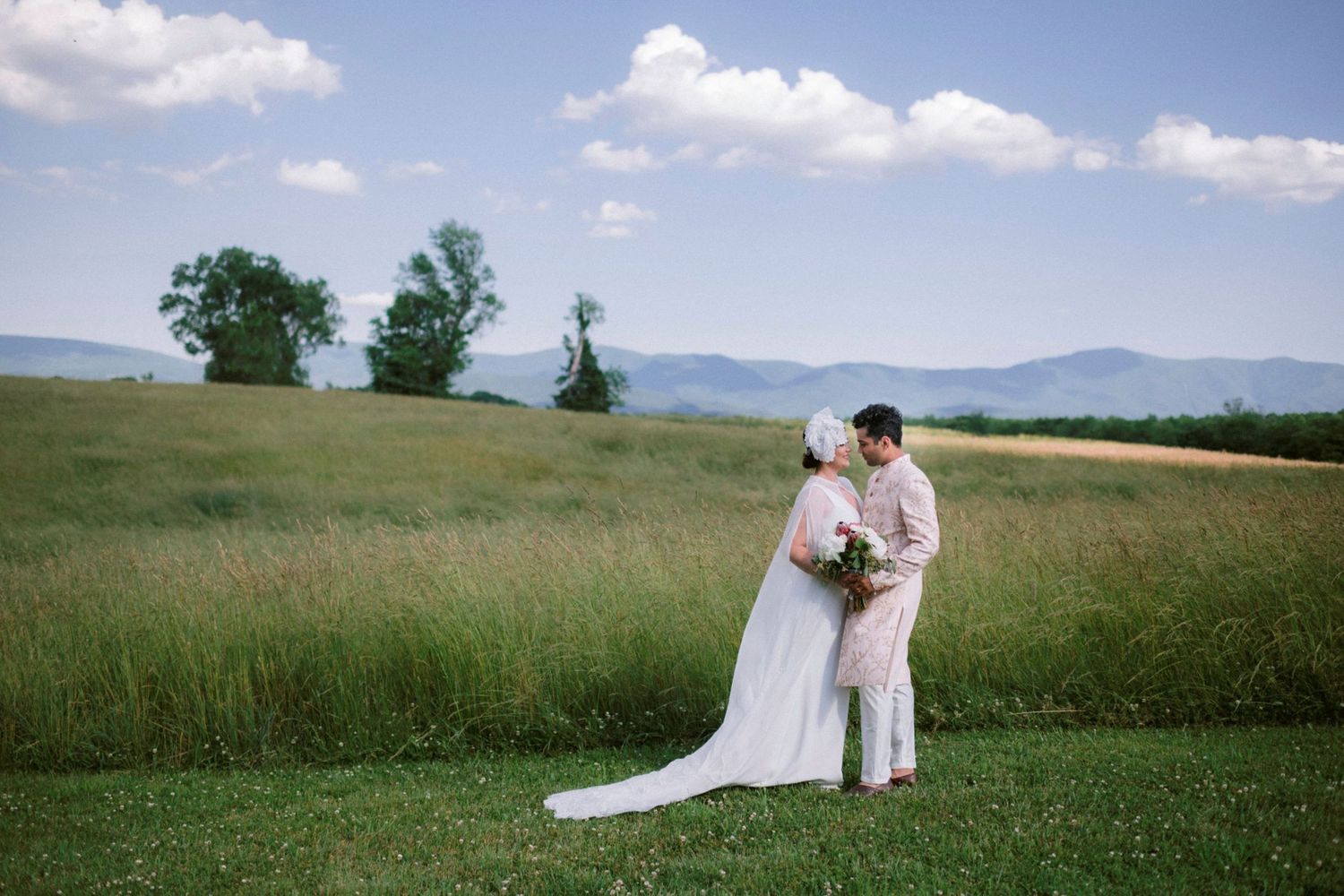 A couple in wedding attire embrace on a hilltop meadow with mountains and blue sky in the background.