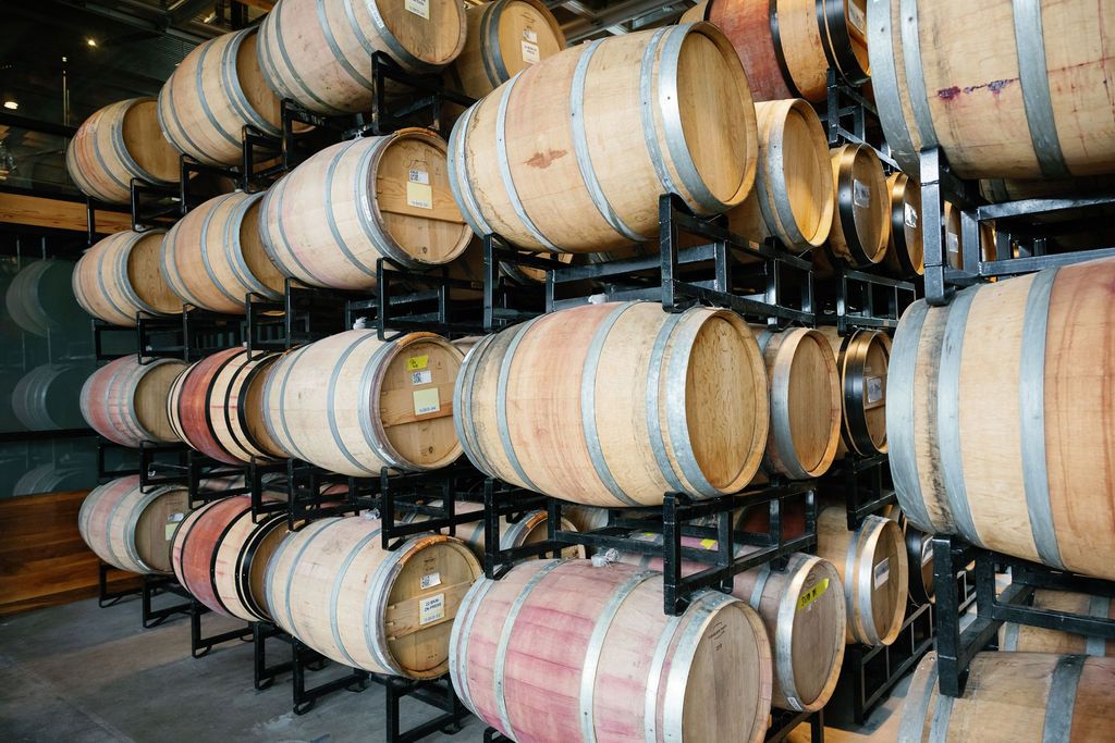 Stacks of wooden wine barrels stored in a cellar aging room with metal support racks.