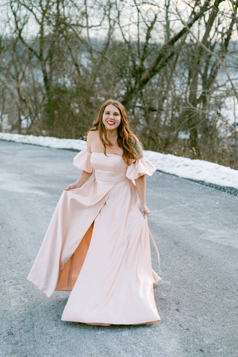 Person in flowing pink dress twirls gracefully on snowy winter road surrounded by bare trees.